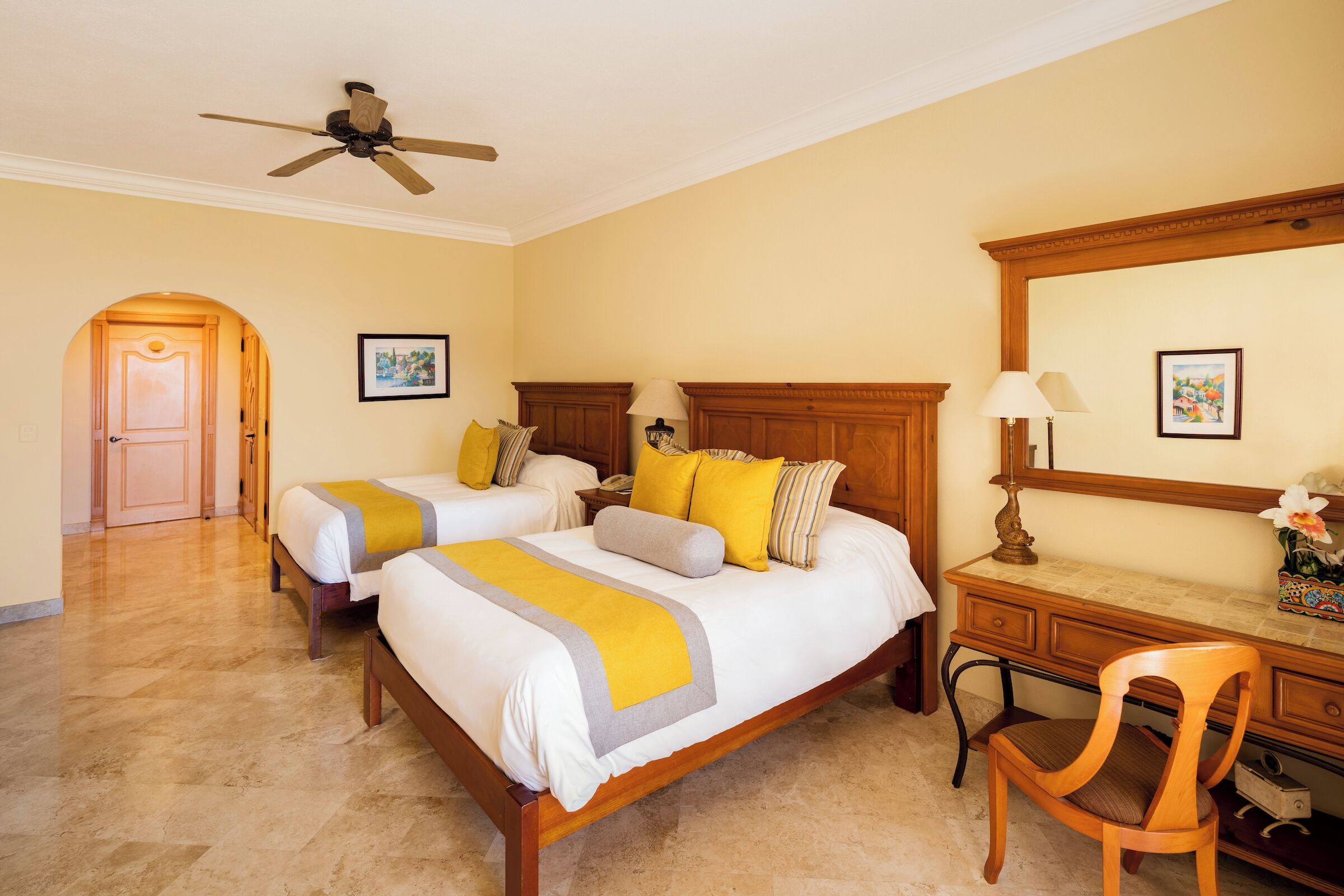 A hotel room with two beds, a desk, a chair, a ceiling fan, and a door leading to another room.