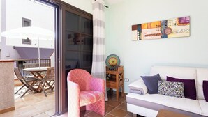 Ein Wohnzimmer mit einem weißen Sofa, einem gemusterten Sessel, einem kleinen Tisch mit einer Dekorplatte und Blick auf eine Terrassenanlage mit Tisch und Stühlen.