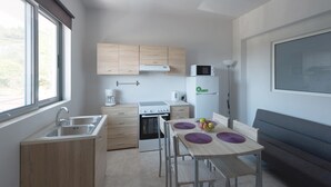A compact kitchen with a sink, stove, and microwave. There is a dining table with purple placemats and a bowl of fruit. - Paulsia Apartments (Rhodes)