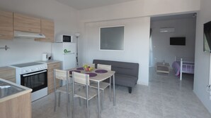 A modern kitchen with a stove, microwave, and refrigerator. There is a dining table with chairs and a gray sofa in the living area. - Paulsia Apartments (Rhodes)