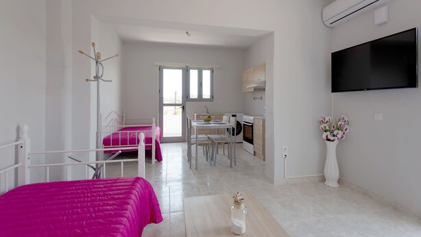 A bedroom with a bed, a bedside table, and a window. There is a kitchenette with a sink, stove, and cabinets. A television is mounted on the wall. - Paulsia Apartments (Rhodes)