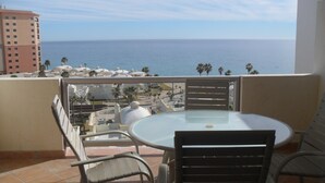Ein Balkon mit rundem Tisch und Stühlen, der Blick auf die Küste mit Gebäuden und Palmen bietet.