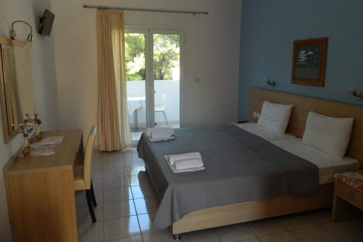 A hotel room with a bed, a desk, a chair, a window with curtains, and a door leading to a balcony.