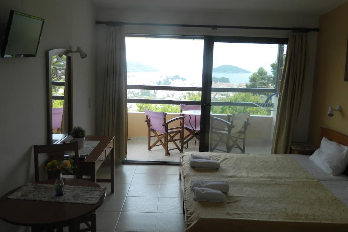A hotel room with a bed, a desk with a chair, a small table with a vase, and a balcony with a table and chairs.