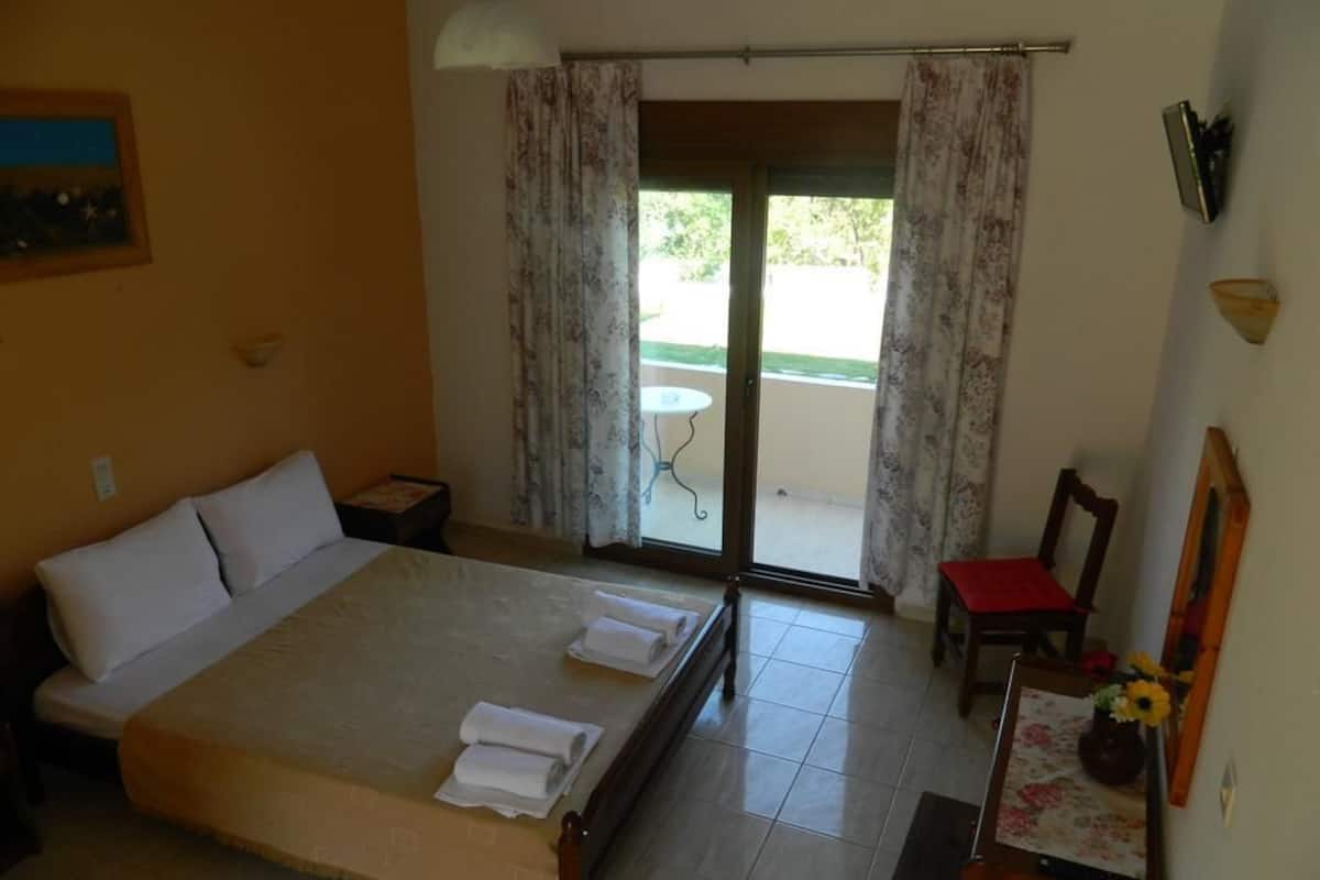 A hotel room with a bed, a desk, a chair, a small table with a vase of flowers, and a sliding glass door leading to an outdoor area.