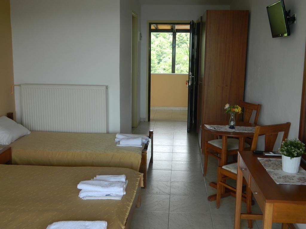 A hotel room with two beds, a dining table with chairs, a TV mounted on the wall, and a balcony with a view of greenery.
