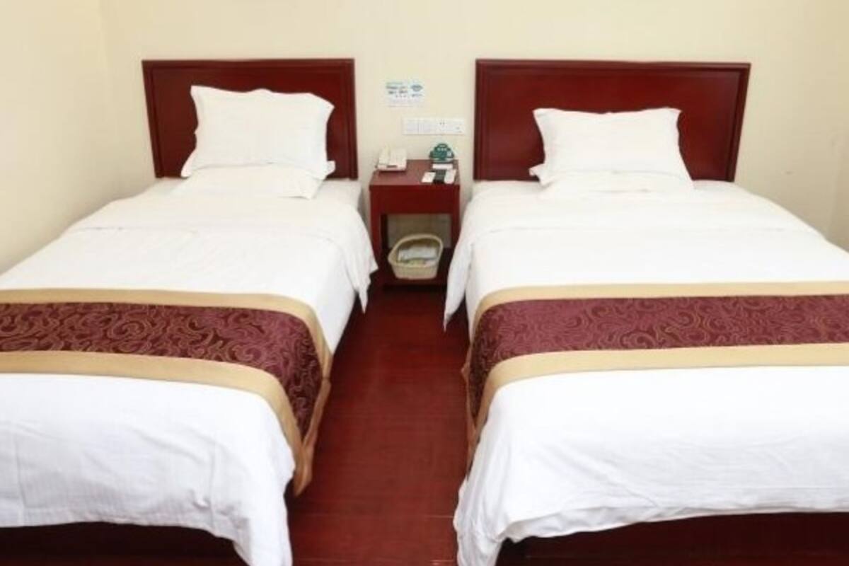 A hotel room with two single beds, each with a wooden headboard and white bedding. There are two wall-mounted paintings, one depicting a landscape and the other a body of water.