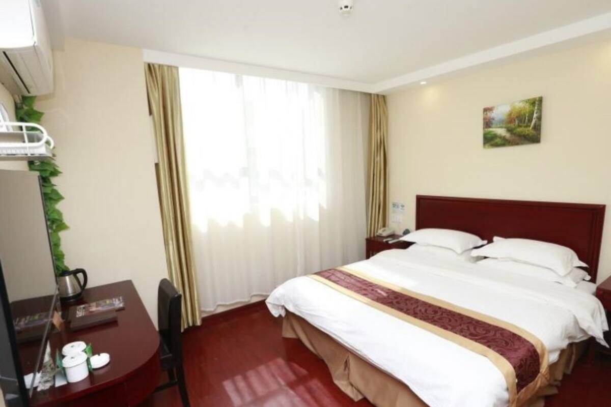 A hotel room with a large bed, a desk with a chair, a small table with a kettle, and a window with curtains.