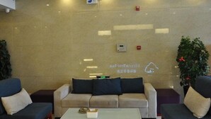 Zona de recepción con un sofá color beige, una mesa de centro y dos sillones. Hay plantas, un panel de control en la pared y un detector de incendios.