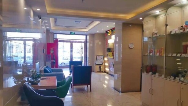 Un vestíbulo de hotel con mostrador de recepción, zona de estar con sillas, vitrina de vidrio y suelo de baldosas.