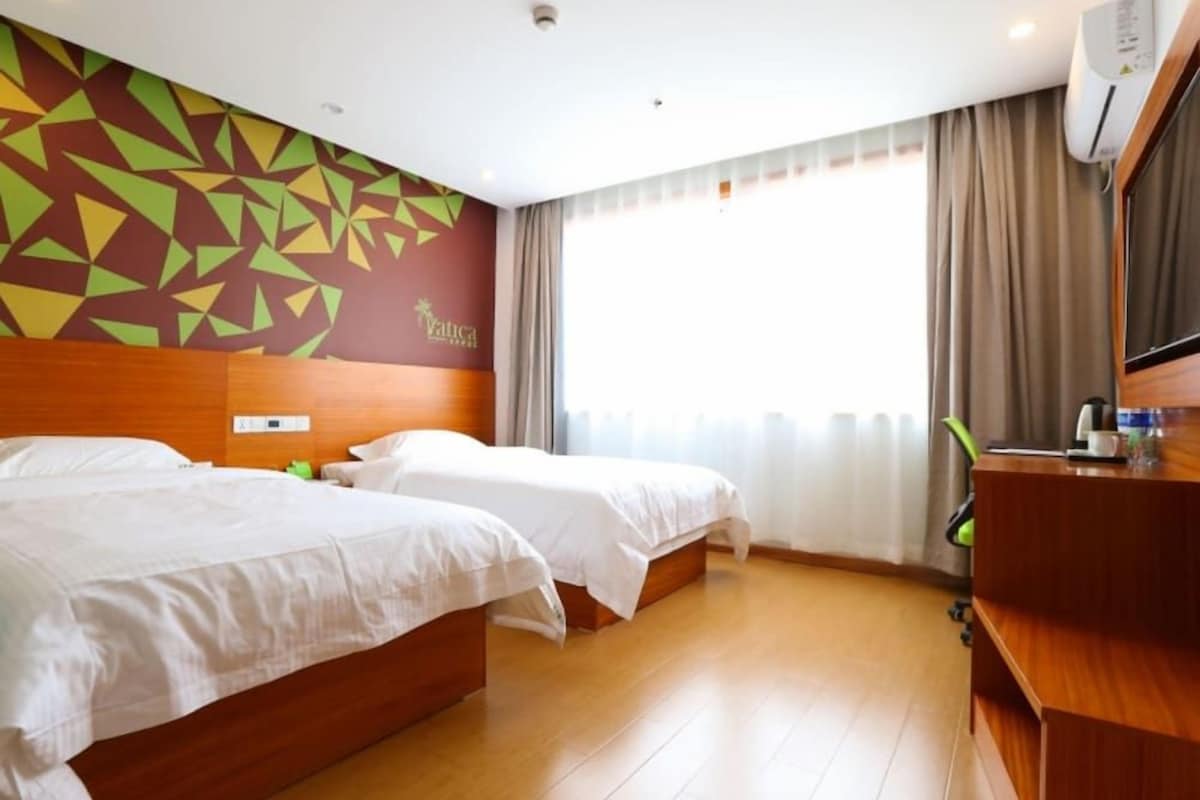 Camera d'albergo con due letti, una testiera in legno, una scrivania con sedia e un'ampia finestra con tende.