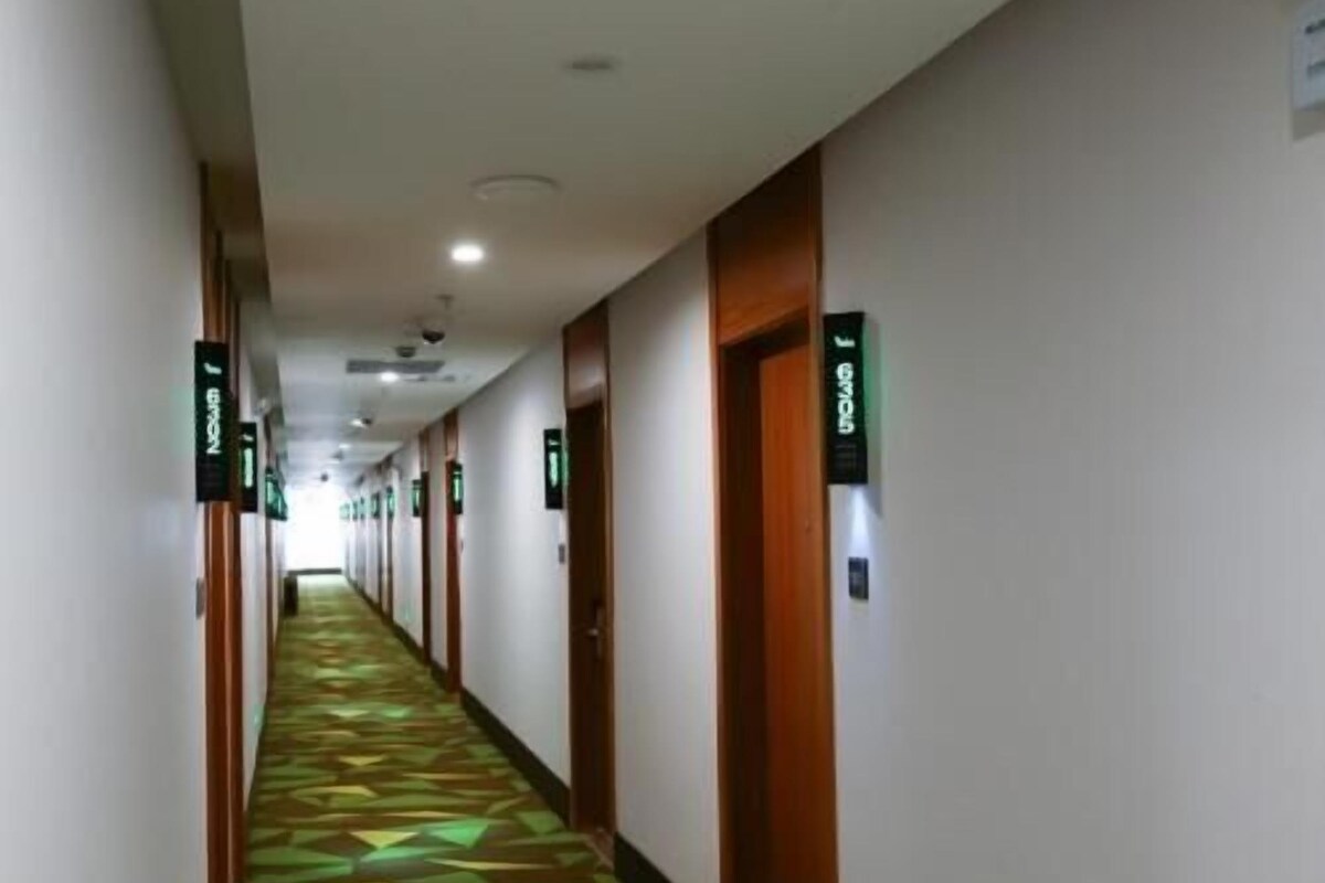 Een hotelgang met deuren aangeduid met groene bordjes, een kamerbreed tapijt en een plafond met inbouwverlichting.