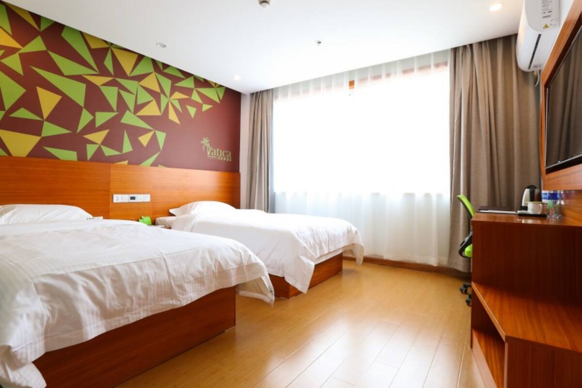 Une chambre d’hôtel avec deux lits, une tête de lit en bois, une fresque murale aux motifs géométriques, une grande fenêtre avec des rideaux et un bureau doté d’un miroir et d’une machine à café.