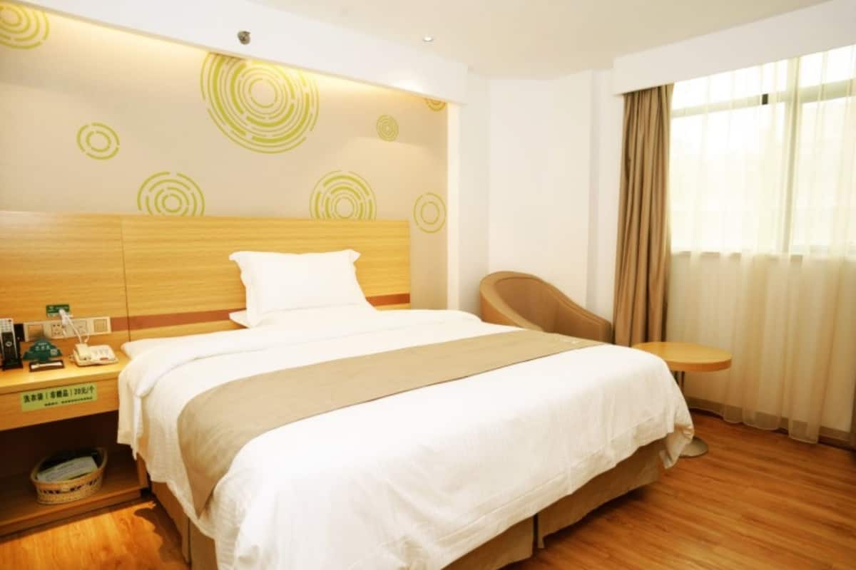 A hotel room with a wooden bed, a bedside table with a lamp, a chair, and a window with curtains.