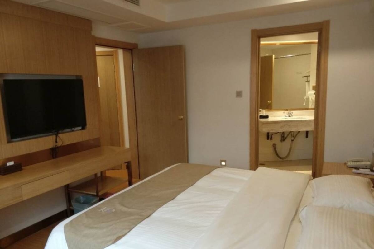 A hotel room with a bed, a television on a wooden stand, a bathroom with a mirror and sink, and a door leading to another room.