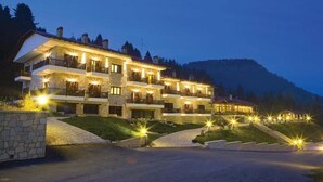 Un immeuble en pierre à plusieurs étages, doté de balcons et illuminé par des éclairages extérieurs, se dressant sur un fond de paysage montagneux.