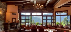 A cozy room with a fireplace, large windows with mountain views, and a chandelier. - Hotel Magiossi (Pyli)
