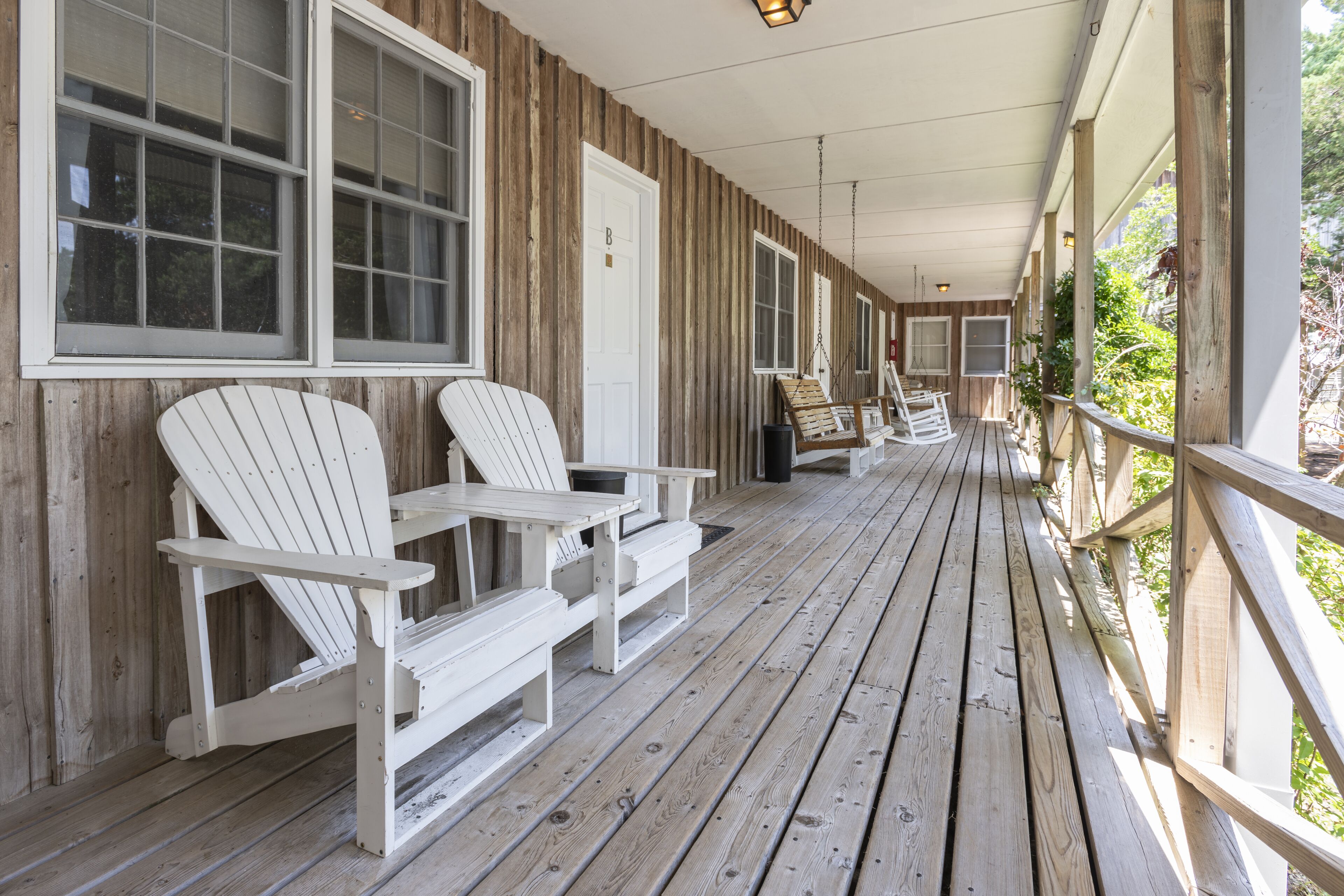 A wooden porch with Adirondack chairs and a swing chair, featuring a wooden railing and large windows.