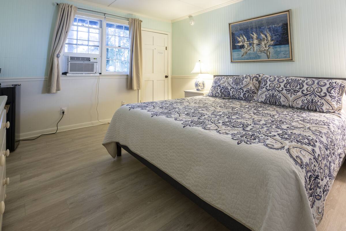 Une chambre à coucher avec un lit, une table de chevet, une lampe, une fenêtre avec des rideaux et un tableau au mur.