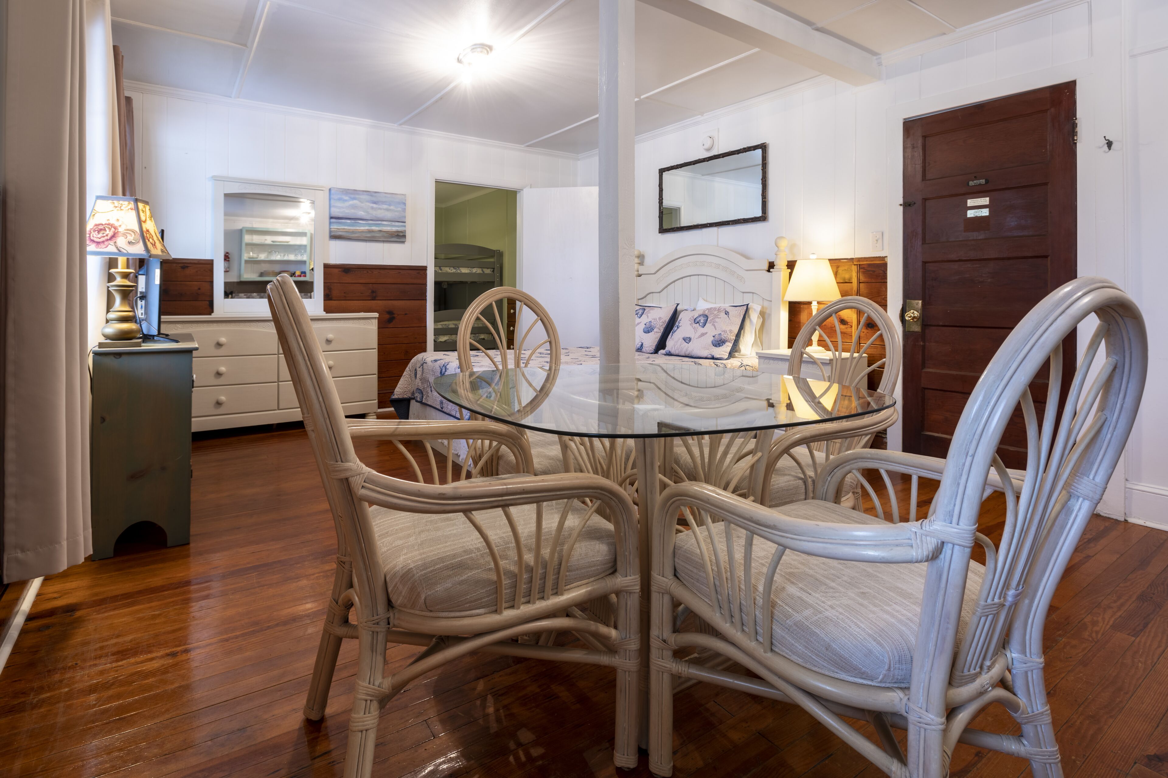 A dining area with a glass-top table surrounded by wicker chairs, a wooden cabinet, and a lamp.