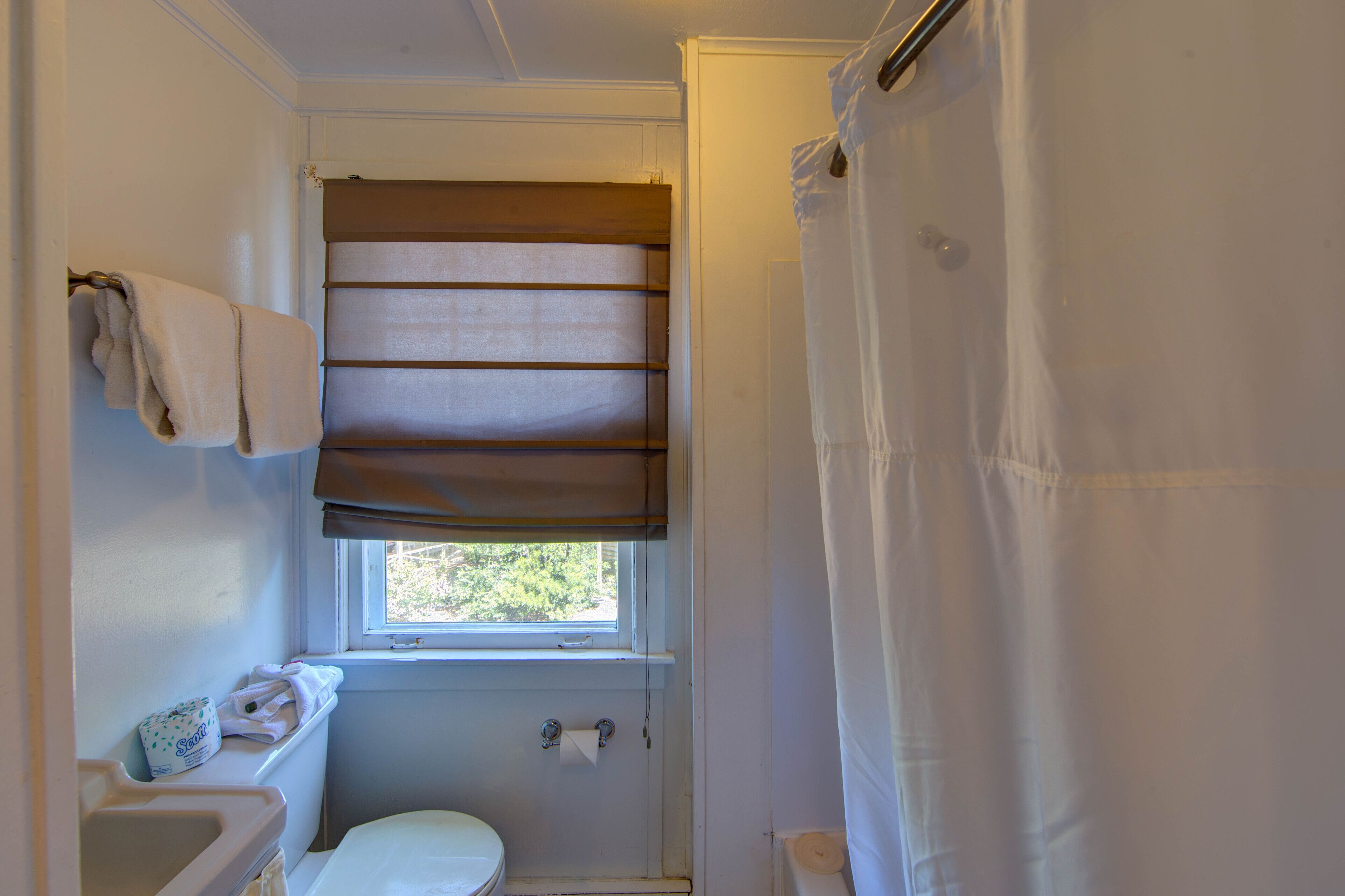 A small bathroom with a toilet, a sink, a window with blinds, and a shower curtain.