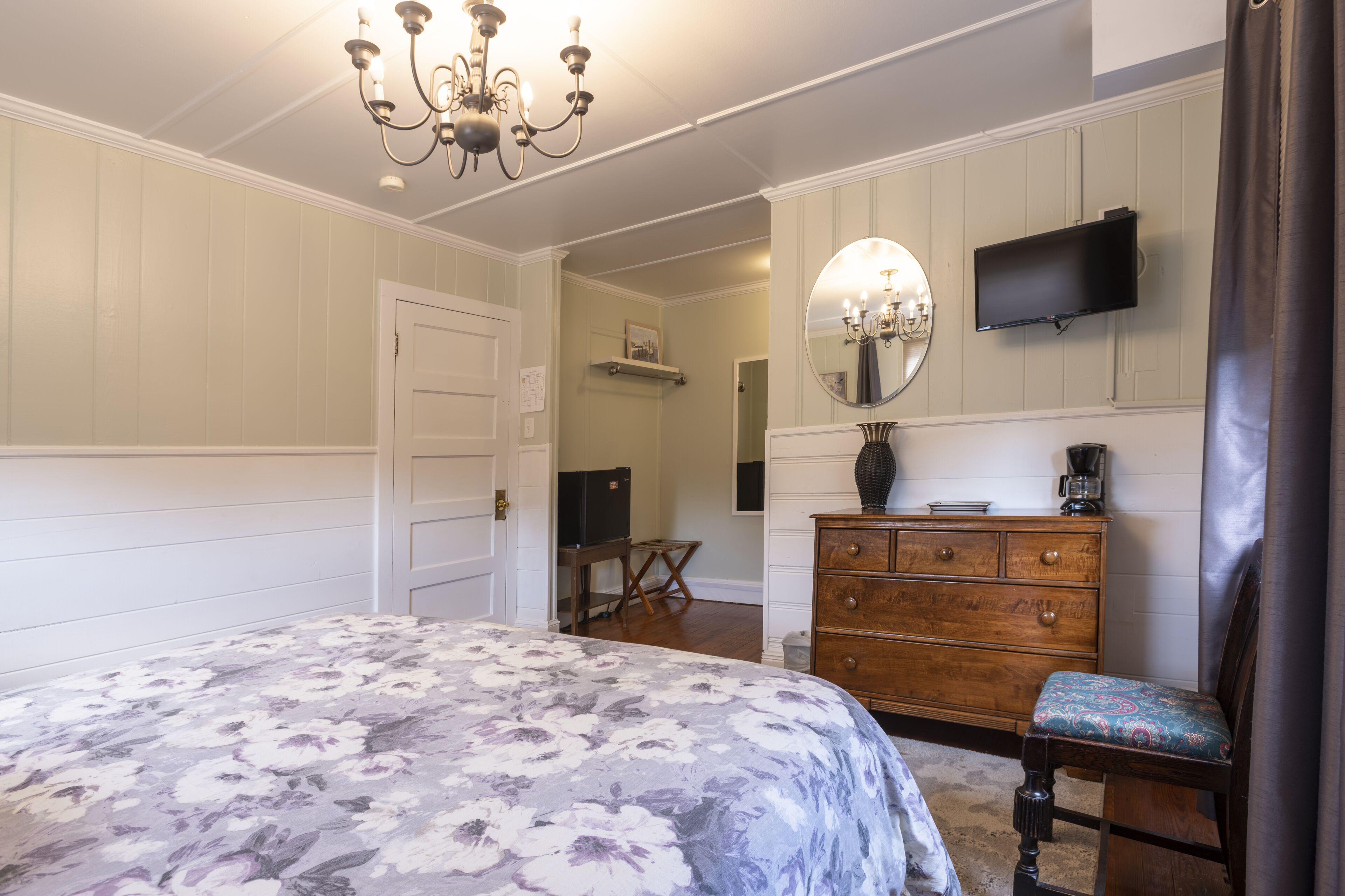 Ein Schlafzimmer mit einer blumigen Tagesdecke, einem Holzkleiderschrank, einem Sessel, einem Fernseher und einer Chandelüster.