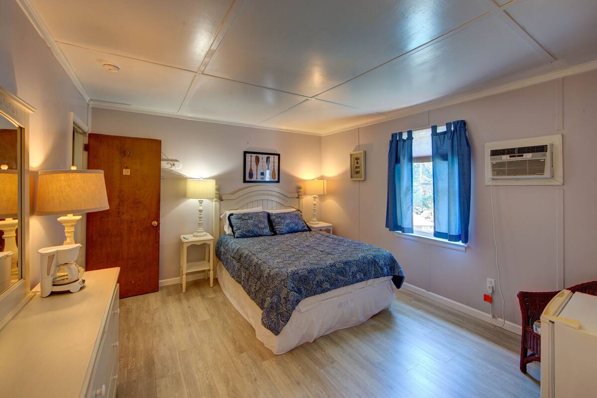 Une chambre à coucher comprenant un lit, des tables de chevet, une commode, un miroir, une porte et une fenêtre avec des rideaux.