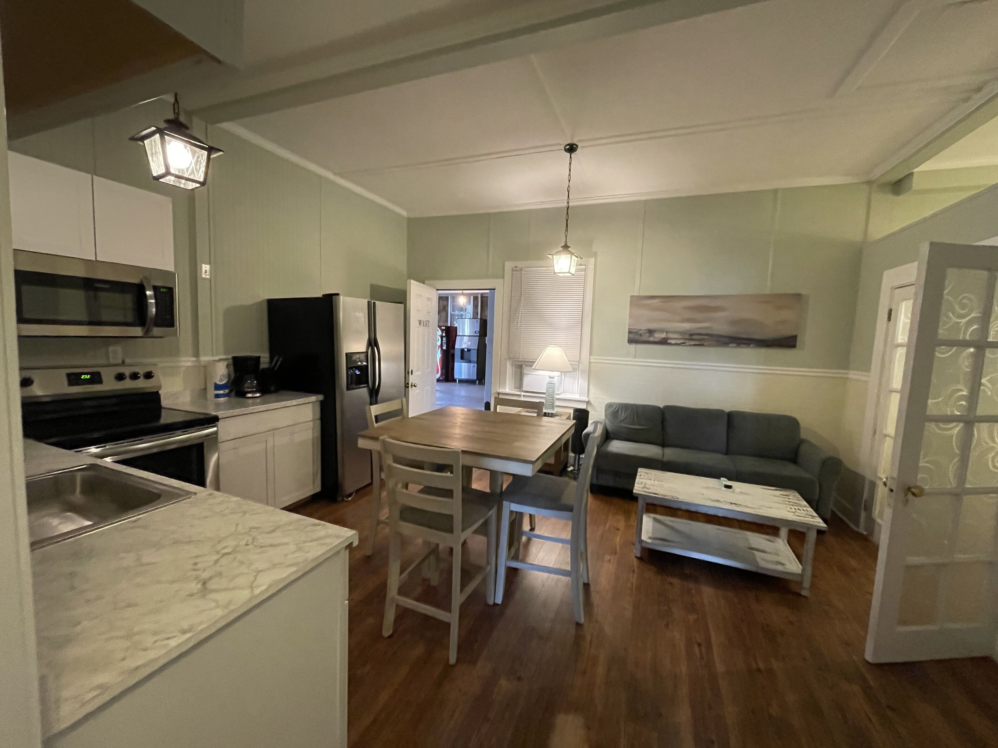 A compact kitchen with a microwave, stove, and refrigerator. There is a dining table with chairs and a sofa in the living area.