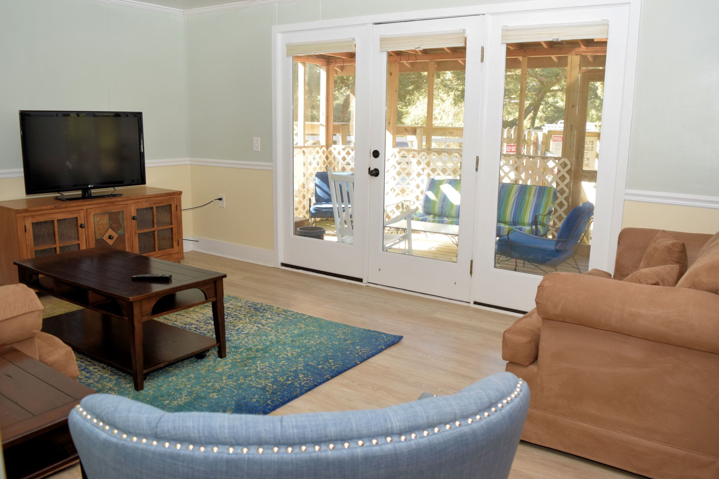 A living room with a TV on a wooden cabinet, a blue armchair, a brown sofa, and a sliding glass door leading to an outdoor patio area.