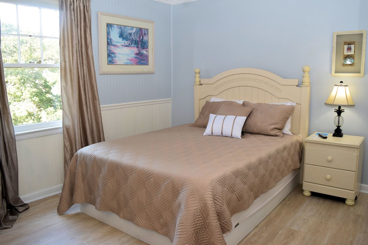 Un lit bien fait, avec une couvre-lit beige et des oreillers, une table de chevet avec une lampe, et un tableau encadré accroché au mur.