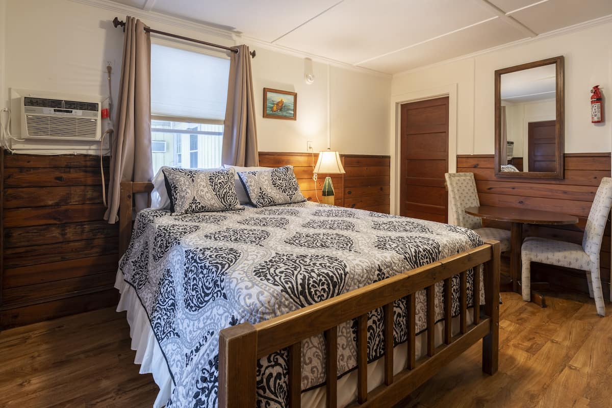 Une chambre avec un lit en bois, une table de chevet avec une lampe, une chaise, une fenêtre avec des rideaux et un climatiseur fixé au mur.