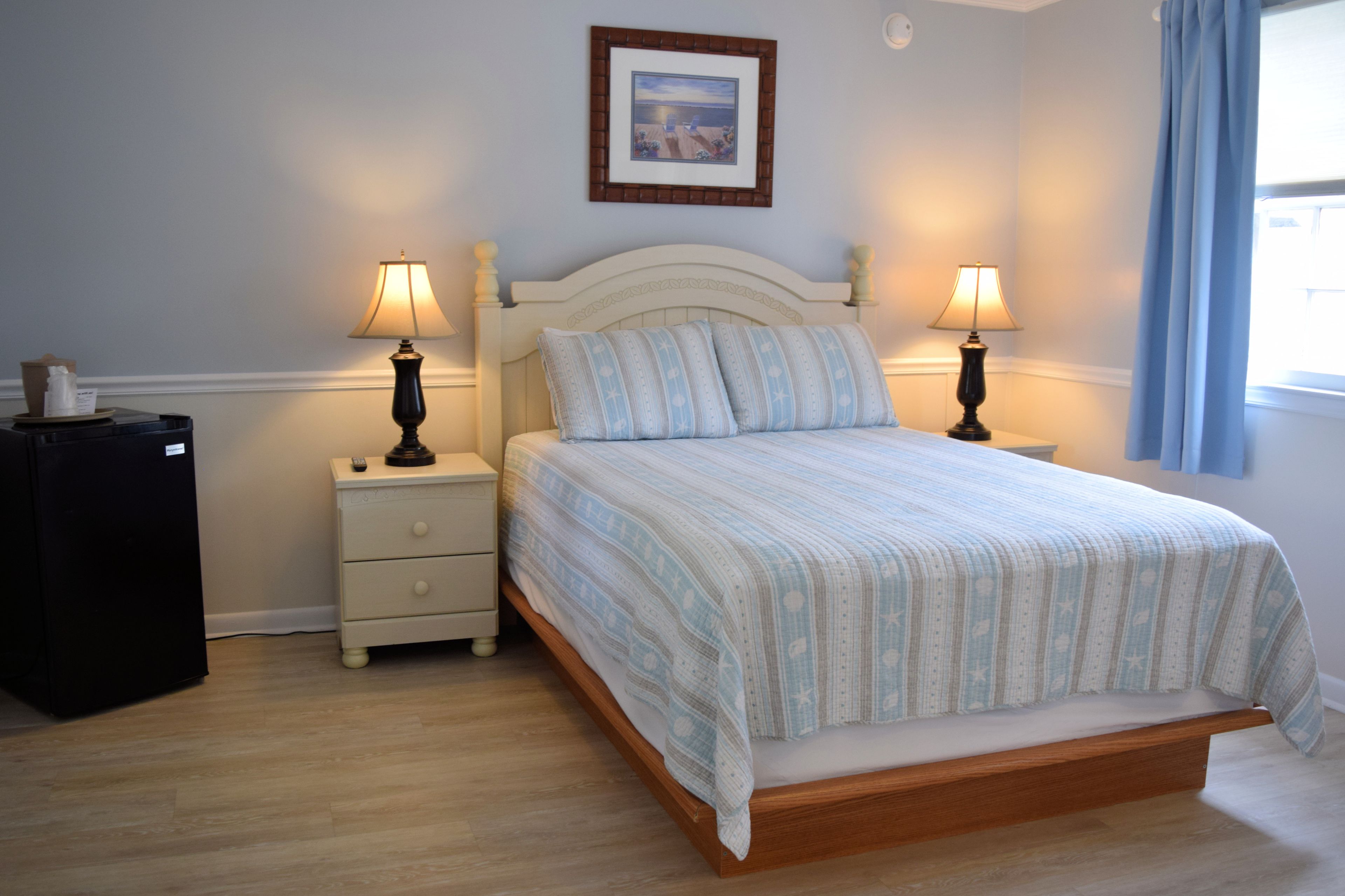 A neatly made bed with a patterned bedspread, two bedside lamps, a small bedside table with a lamp, and a framed picture on the wall.