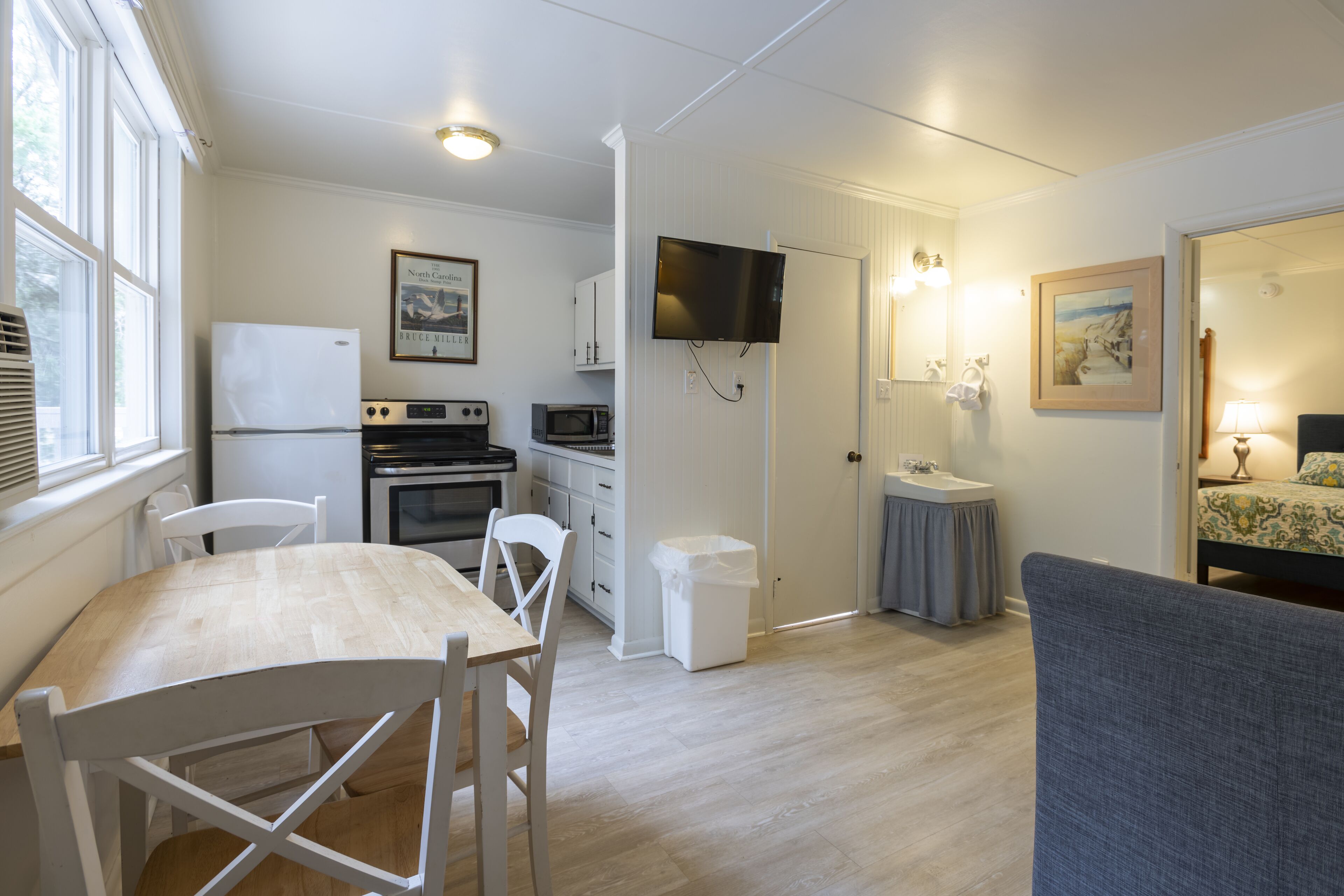 A compact kitchen with a refrigerator, microwave, and stove, a dining area with a table and chairs, and a bedroom with a bed and nightstand.