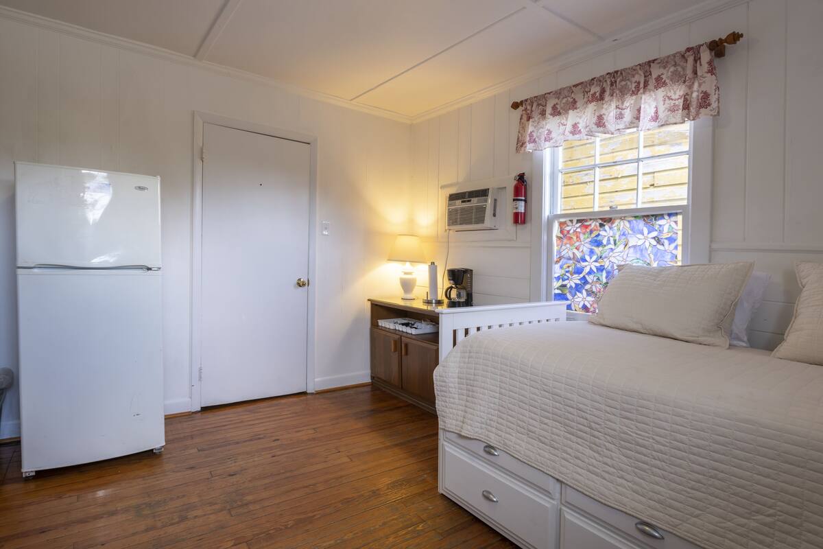 Une chambre à coucher comprenant un lit, une table de chevet, une lampe, une fenêtre agrémentée d’une lambrequin à motifs floraux et un réfrigérateur.