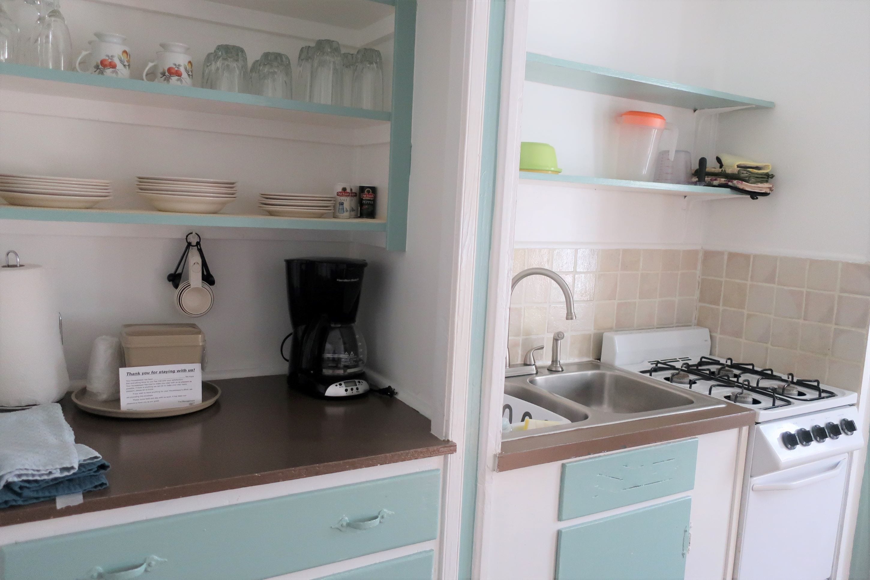 A kitchen with a stove, sink, and shelves with dishes and kitchenware.