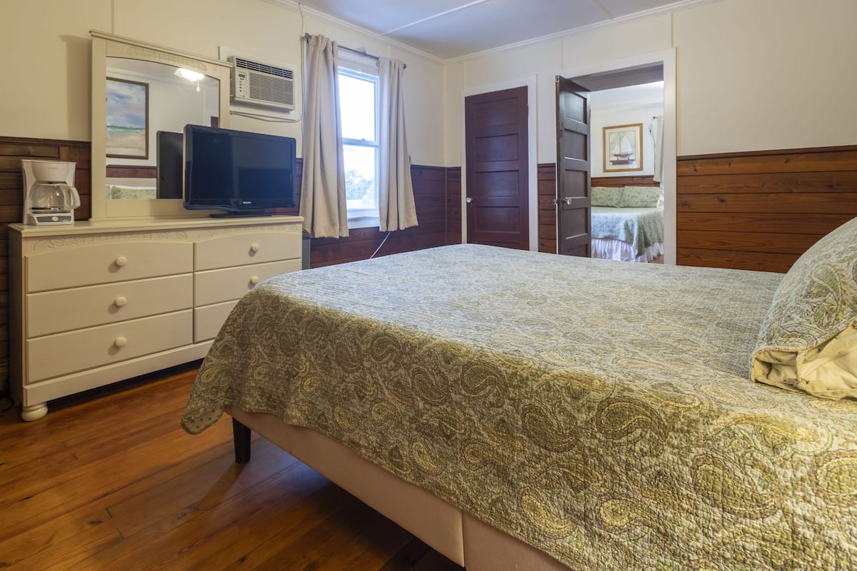Une chambre à coucher avec un lit, une commode, un téléviseur et une fenêtre avec des rideaux.