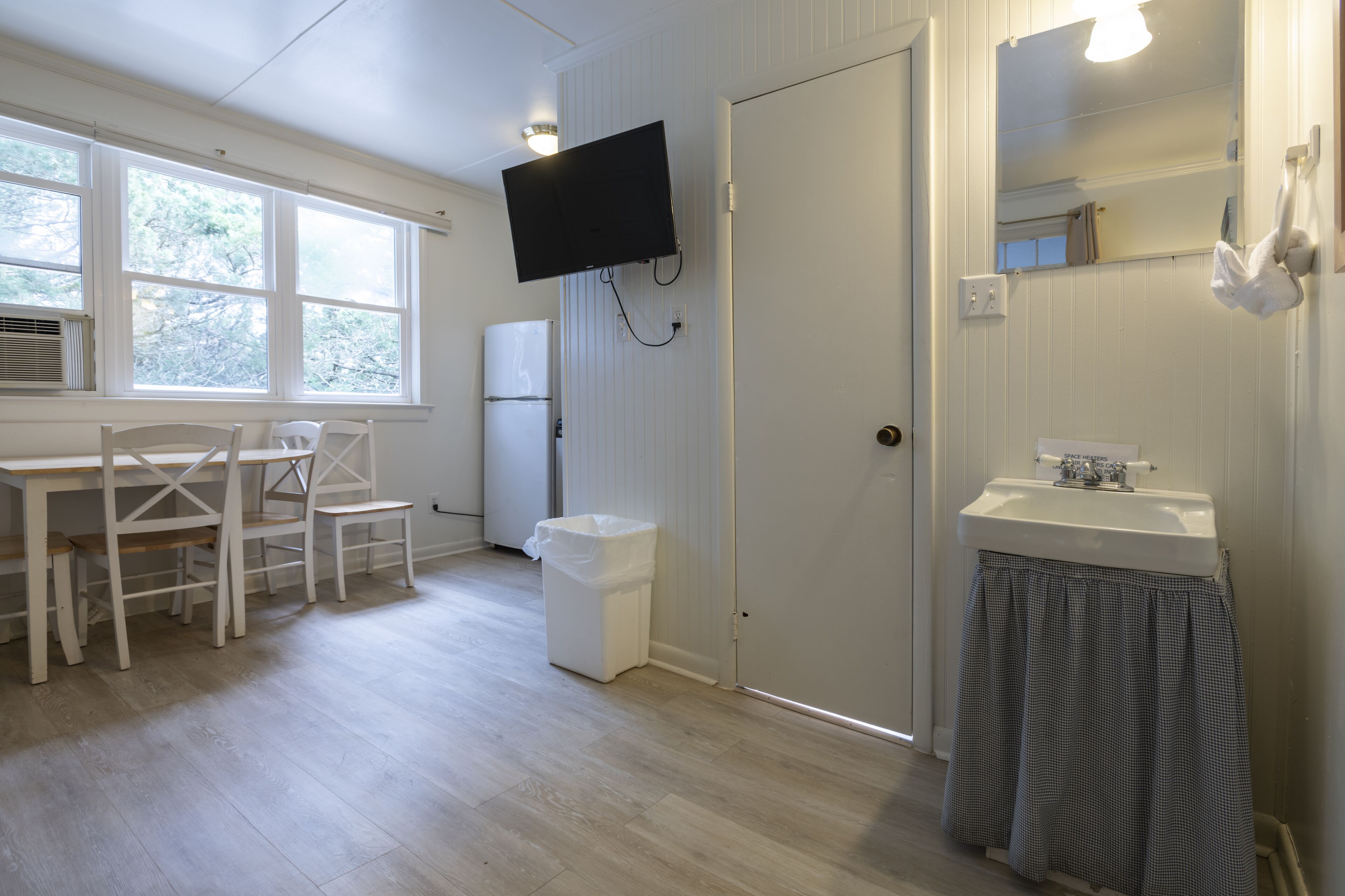 Une pièce avec une table et des chaises blanches, un lavabo blanc, un miroir et un téléviseur fixé au mur.