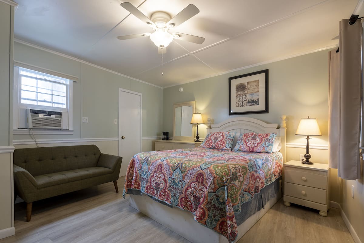 Une chambre à coucher comprenant un lit, une table de chevet, une lampe, un ventilateur de plafond, une fenêtre avec des stores et un canapé.