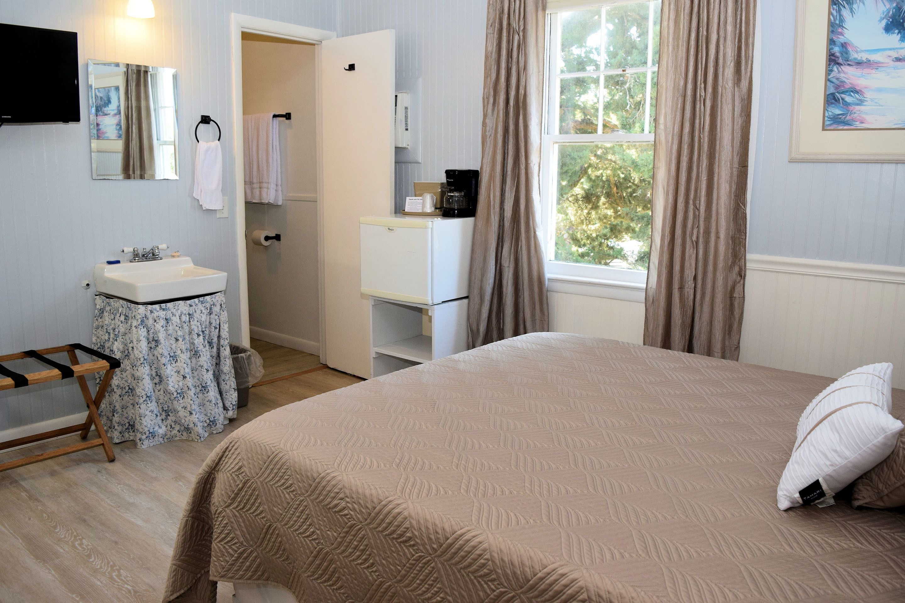 A bedroom with a bed, a nightstand, a dresser, a mirror, a window with curtains, and a bathroom with a sink and towel rack.