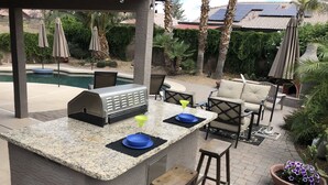 An outdoor bar area with a built-in grill, bar stools, and a pool in the background.