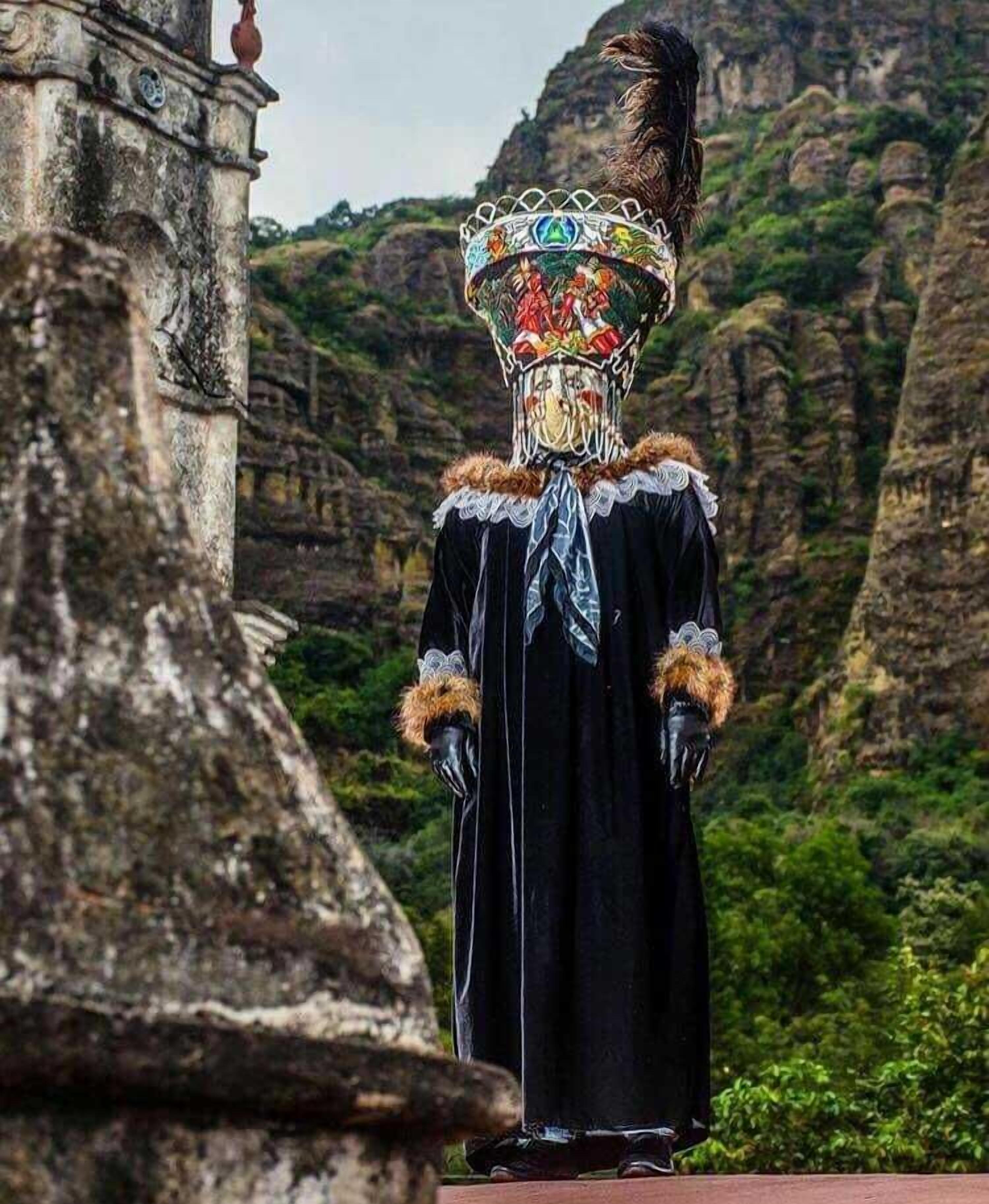 Una persona con túnica negra y tocado decorativo, de pie frente a una construcción de piedra con montañas al fondo.