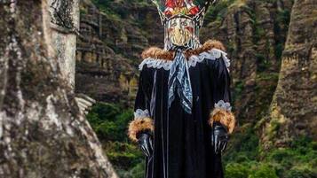Una persona con túnica negra y tocado decorativo, de pie frente a una construcción de piedra con montañas al fondo.