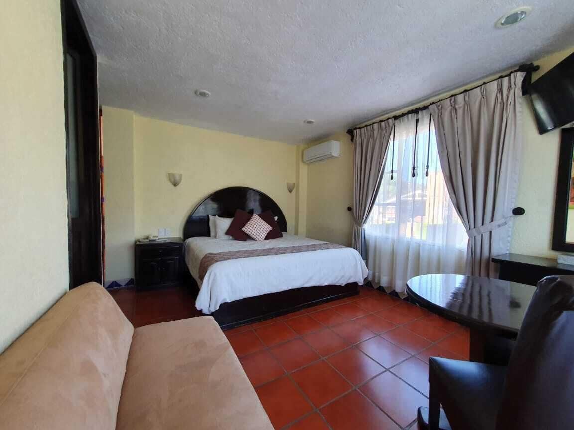 Una habitación de hotel con cama, mesita de noche, una silla, una mesa redonda y una ventana con cortinas.