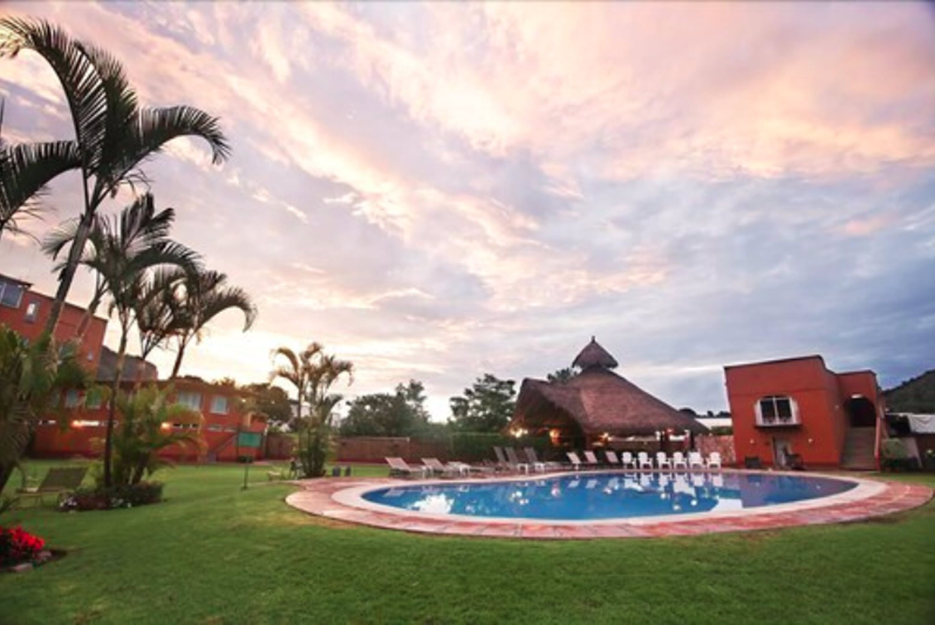 A swimming pool surrounded by a lawn with lounge chairs, a thatched-roof building, and palm trees.