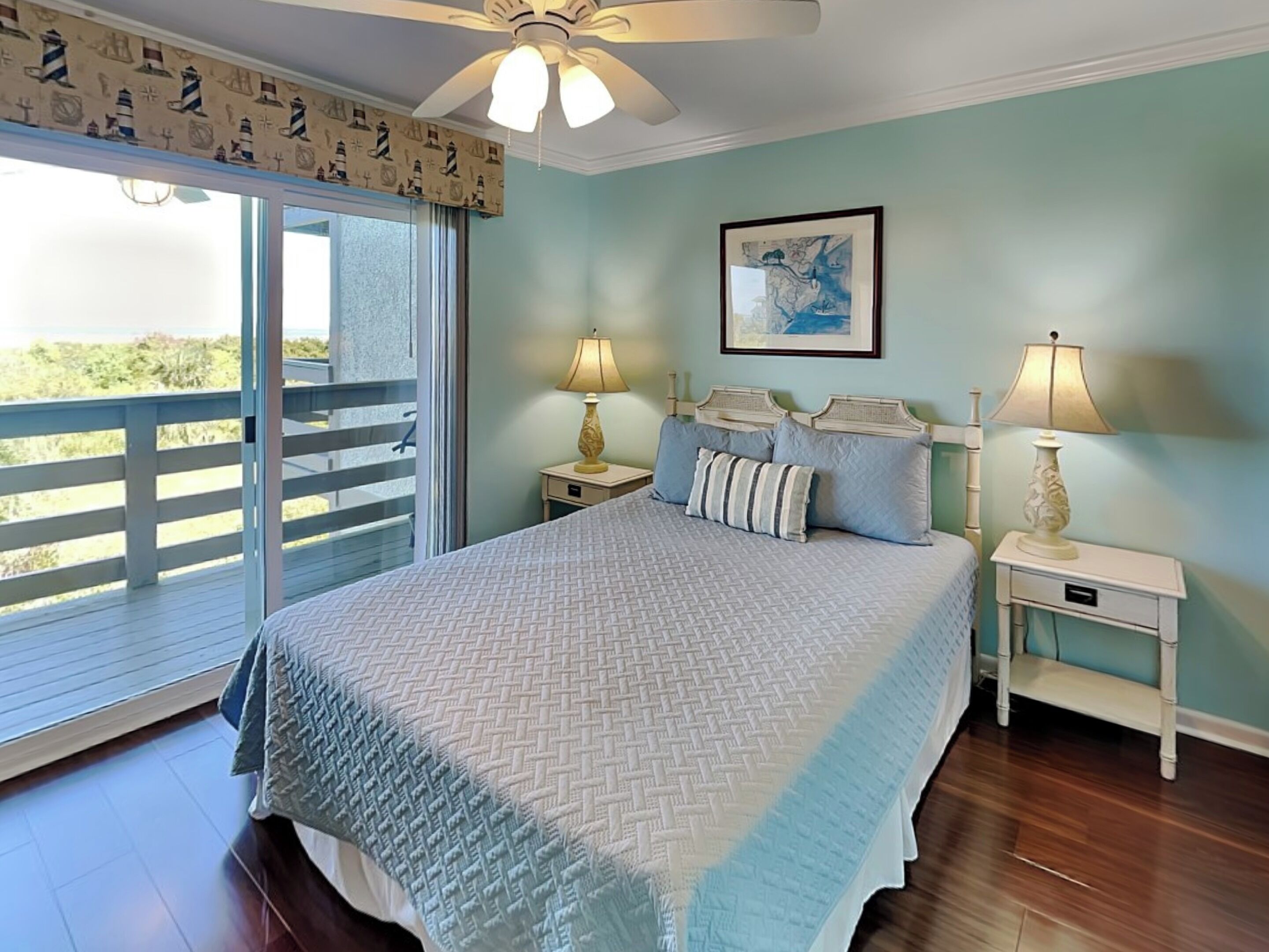 A bedroom with a large bed, two bedside tables with lamps, a ceiling fan, a window with a view, and a framed picture on the wall.