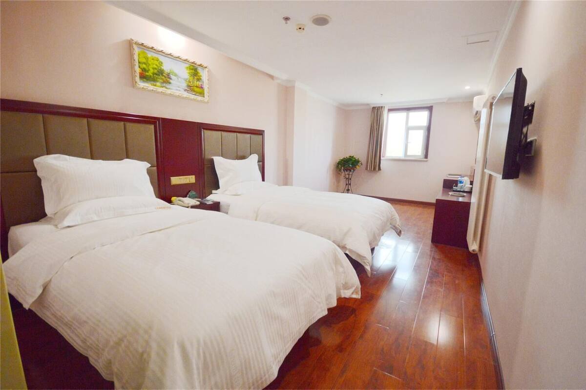 Ein Hotelzimmer mit zwei Betten, einem Holz-Kopfende, einem kleinen Tisch mit Pflanze, einem Fenster mit Vorhängen und einem an der Wand montierten Fernseher.