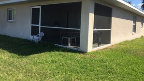 Ein eingeschossiges Gebäude mit einem großen Glastür-Eingang, einer kleinen Terrasse mit einem weißen Kunststoffstuhl und einer Rasenfläche.
