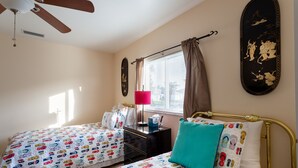 Ein Schlafzimmer mit zwei Betten, einem Deckenventilator, einem Fenster mit Vorhängen, einem Nachttisch mit Lampe und Wandbildern.