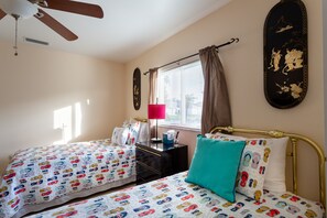 A bedroom with two beds, a ceiling fan, a window with curtains, a bedside table with a lamp, and wall art.