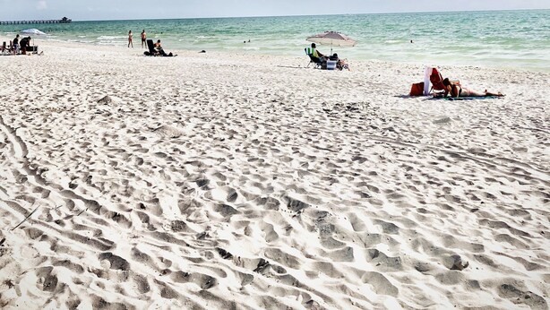 Ein sandiger Strand mit Fußabdrücken, ein Steg im Hintergrund und einige entspannte Personen.